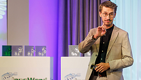 Ein Mann in einem beigen Anzug steht auf einer Bühne und spricht. Er hält einen kleinen Gegenstand in der Hand und hebt den Zeigefinger, als ob er um Aufmerksamkeit bittet. Im Hintergrund sind mehrfarbige Auszeichnungen sichtbar.