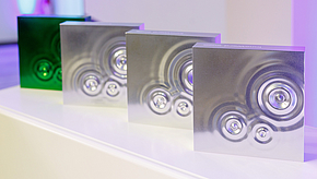 Vier quadratische Objekte mit glänzenden Oberflächen in verschiedenen Farbtönen (eins grün, drei silber) stehen auf einer weißen Fläche. Jedes Objekt zeigt kreisförmige Vertiefungen, die ein strukturiertes Design erzeugen. Die Anordnung betont das moderne und stilvolle Erscheinungsbild der Objekte.