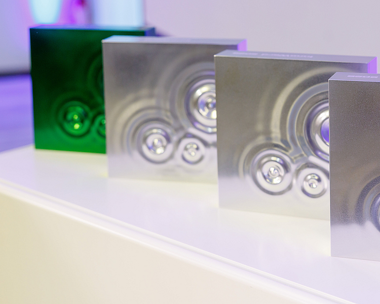 Vier quadratische Objekte mit glänzenden Oberflächen in verschiedenen Farbtönen (eins grün, drei silber) stehen auf einer weißen Fläche. Jedes Objekt zeigt kreisförmige Vertiefungen, die ein strukturiertes Design erzeugen. Die Anordnung betont das moderne und stilvolle Erscheinungsbild der Objekte.