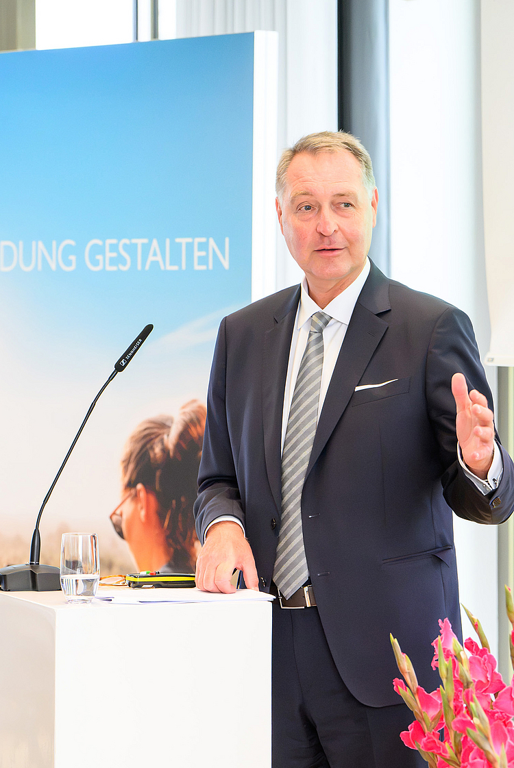 Ein Redner steht an einem Podium und spricht zu einem Publikum. Im Hintergrund ist ein Banner mit dem Text "Vernetzt Bildung Gestalten" und ein Bild von Personen in einer natürlichen Umgebung. Auf dem Podium steht ein Wasserglas. Die Atmosphäre wirkt professionell und inspirierend.
