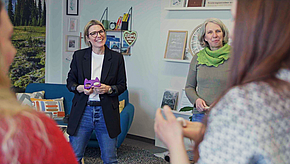 Eine Gruppe von Frauen steht zusammen in einem Raum. Eine Frau mit Brille und schwarzer Blazer lächelt und hält ein lila Objekt in der Hand. Eine andere Frau im Hintergrund trägt einen grünen Schal. Der Raum ist freundlich und einladend gestaltet.