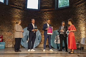 Bei einer Preisverleihung stehen vier Personen auf einer Bühne in einem historischen Raum. Eine Frau in einer gestreiften Jacke, ein Mann in Blau mit einer Auszeichnung, ein weiterer Mann in Anzug und eine Frau in Rot übergeben ihm eine Urkunde.