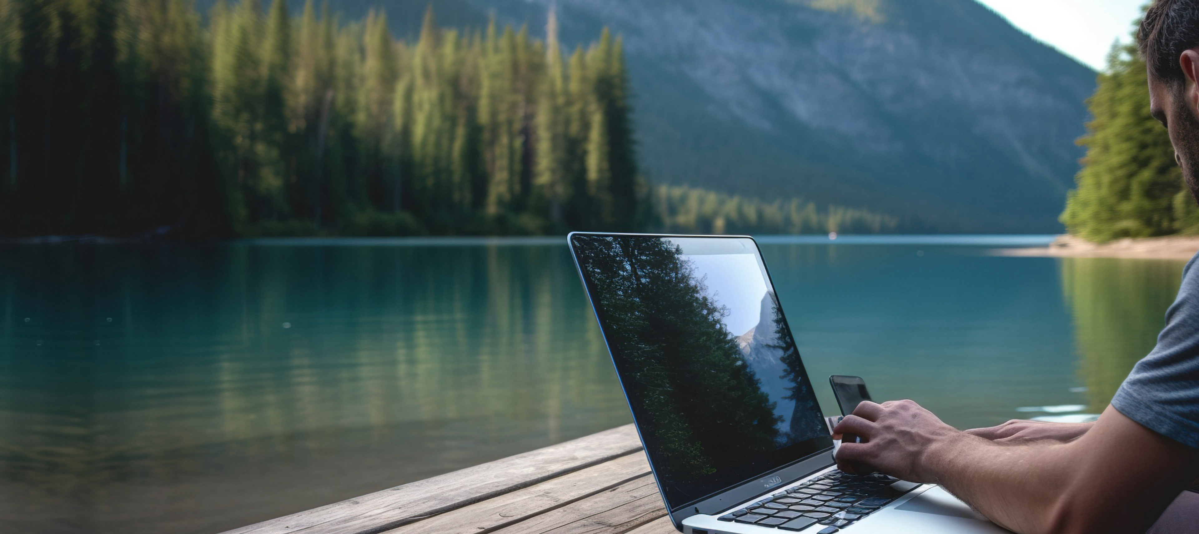 Ein Mann arbeitet an einem Laptop auf einem Holzsteg am Ufer eines ruhigen Sees, umgeben von hohen Bäumen und Bergen im Hintergrund. Das Wasser spiegelt die Landschaft wider, wodurch eine friedliche Atmosphäre entsteht.