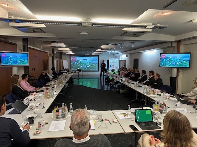 Ein Konferenzraum mit einer langen Tafel, an der mehrere Personen sitzen. An den Wänden sind zwei Bildschirme, auf denen Präsentationen gezeigt werden. Eine Person steht vorne und spricht zu den Anwesenden. Der Raum ist gut beleuchtet.