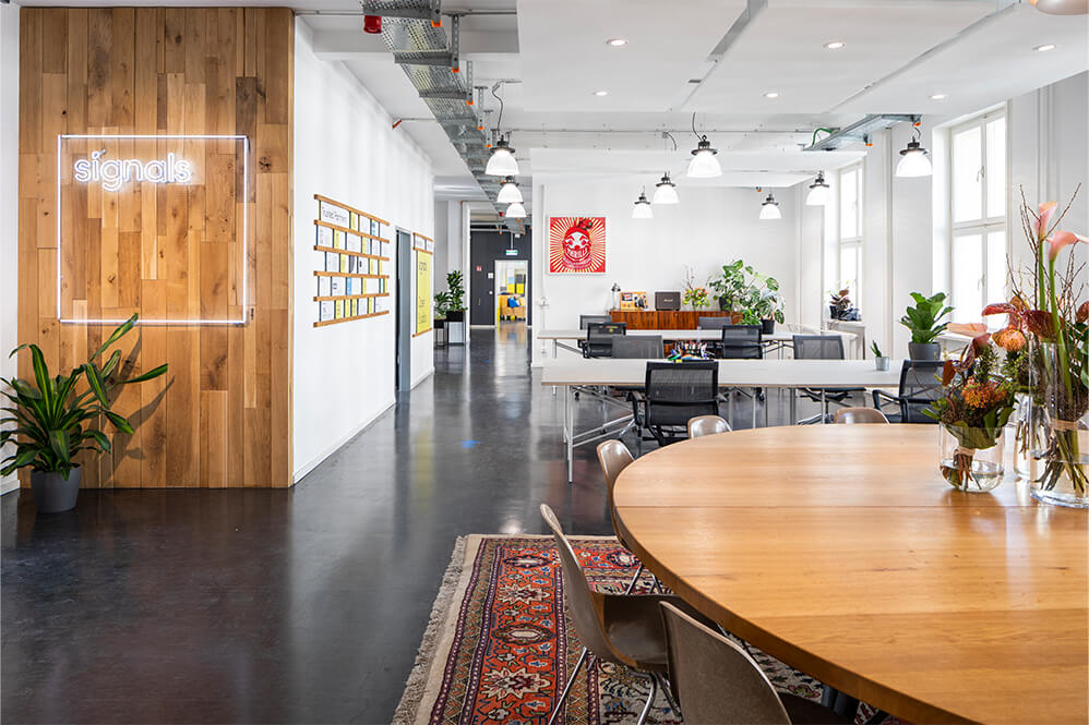 Ein moderner Büroraum mit Holz- und Pflanzenelementen. Links steht ein runder Tisch mit Stühlen, während im Hintergrund mehrere Arbeitsplätze und Pflanzen sichtbar sind. Eine beleuchtete Wand mit dem Schriftzug „signalk“ ist im Eingangsbereich. Helle Beleuchtung und offene Raumgestaltung schaffen eine einladende Atmosphäre.