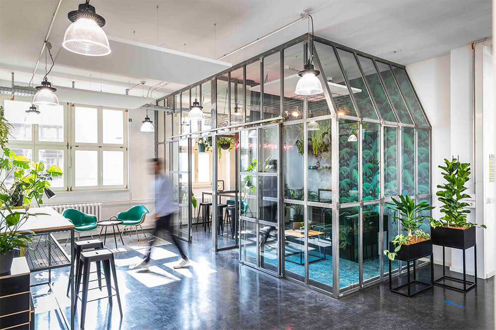 Das Bild zeigt ein modernes Büro mit einer großen Glasstruktur, die als Besprechungsraum genutzt wird. Grünpflanzen sind in und um den Raum platziert, und der Boden ist dunkel. Eine Person bewegt sich durch den Raum. Natürliches Licht strömt durch große Fenster.
