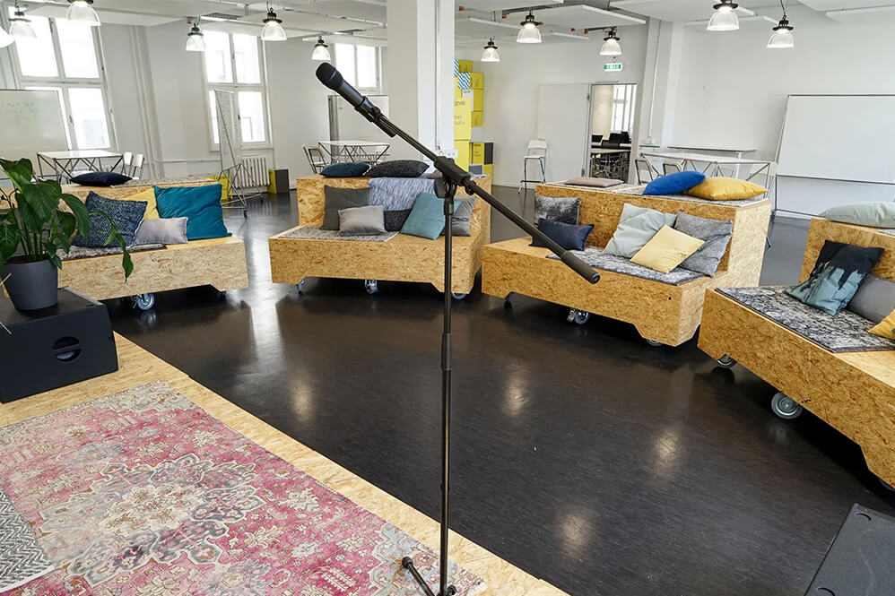 Ein gemütlicher Raum mit modularen Sofas in verschiedenen Farben, die um einen Teppich gruppiert sind. In der Mitte steht ein Mikrofon auf einem Ständer, was auf eine Präsentation oder einen Vortrag hinweist. Die Wände sind hell und es gibt große Fenster, die Tageslicht hereinlassen.